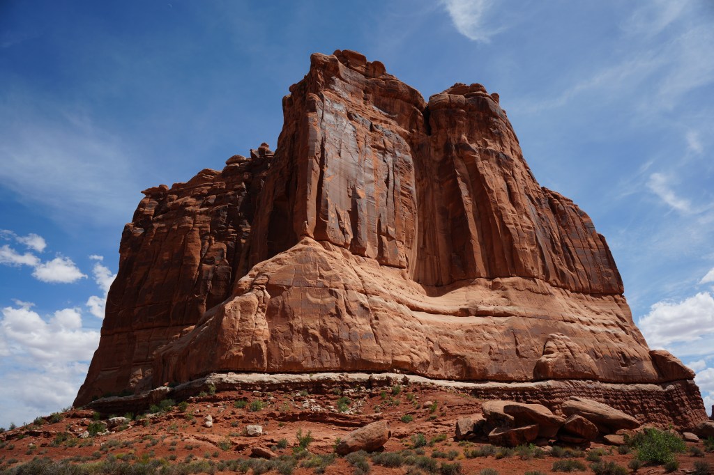 A towering sandstone rock formation against a blue sky with scattered clouds, showcasing the natural beauty of the desert landscape.