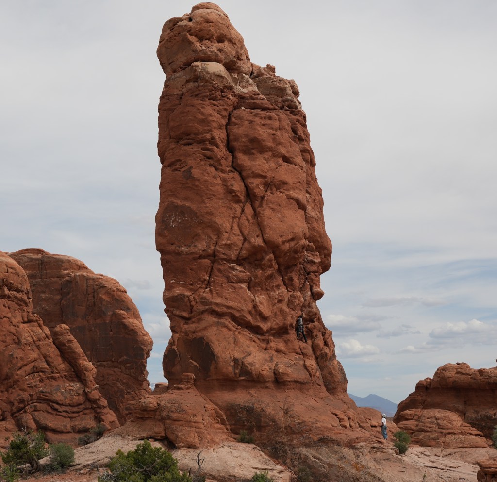 A tall red rock formation in a desert landscape, with two climbers scaling the side and a cloudy sky in the background.