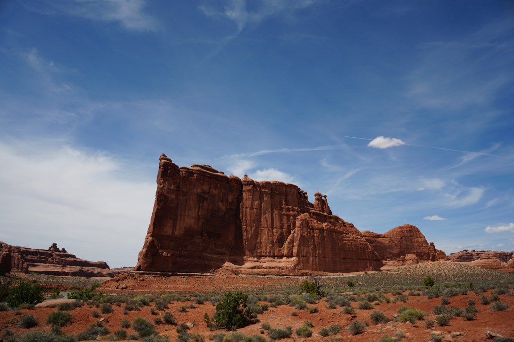 A scenic view of towering red rock formations under a blue sky with scattered clouds, surrounded by desert vegetation.