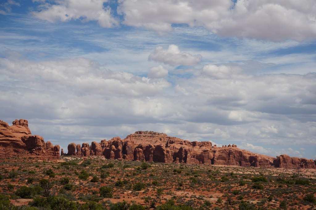 A scenic view of rocky formations under a partly cloudy sky, showcasing reddish-brown cliffs and a sparse landscape with low shrubs.