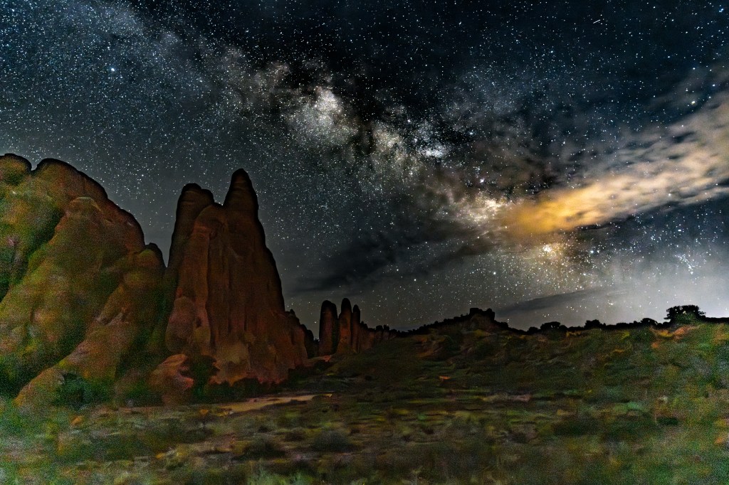 A starry night sky with the Milky Way visible above rugged rock formations and a dark landscape.