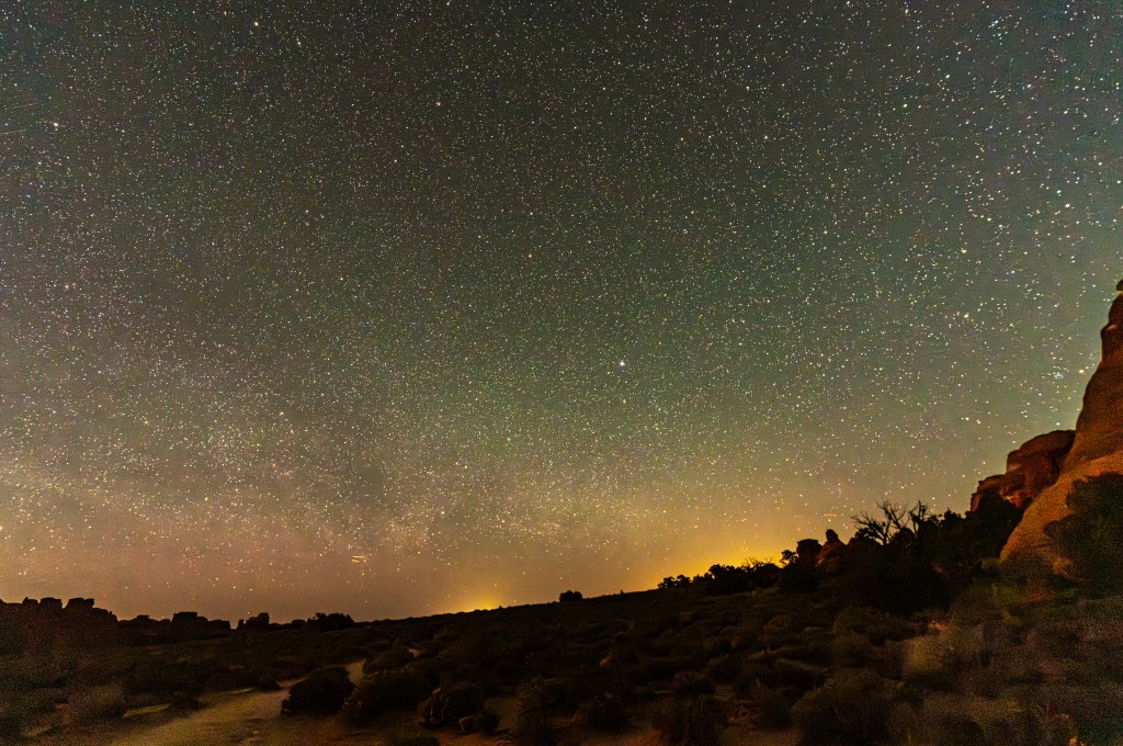 A night sky filled with countless stars, with a gradient of colors towards the horizon. The landscape below features silhouettes of desert terrain and shrubs under the starlit sky.