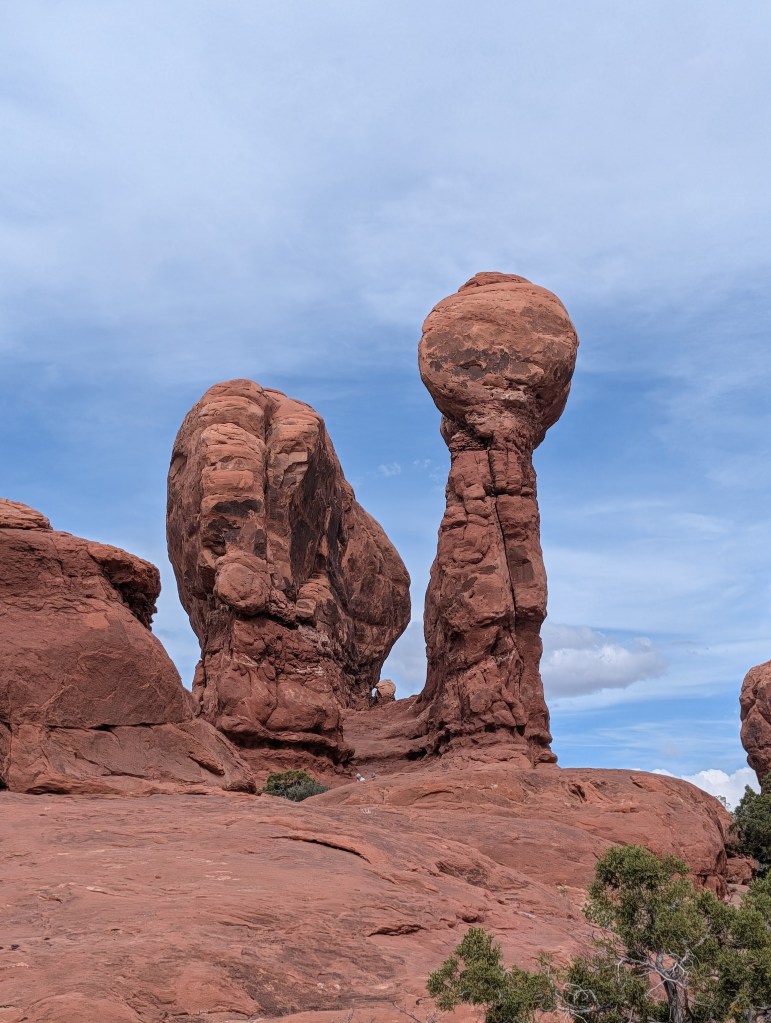 A landscape featuring large red rock formations, including a tall, slender rock topped with a round boulder against a blue sky.