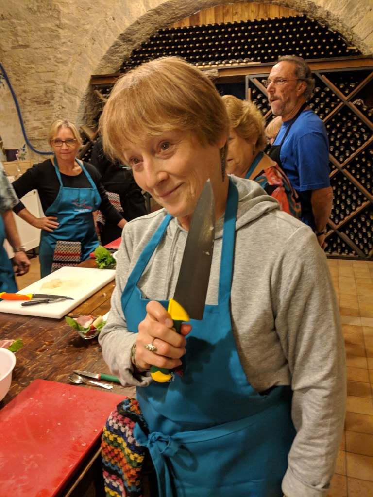 A woman in a blue apron holds a large kitchen knife, smiling playfully in a cooking class setting with other participants in the background.