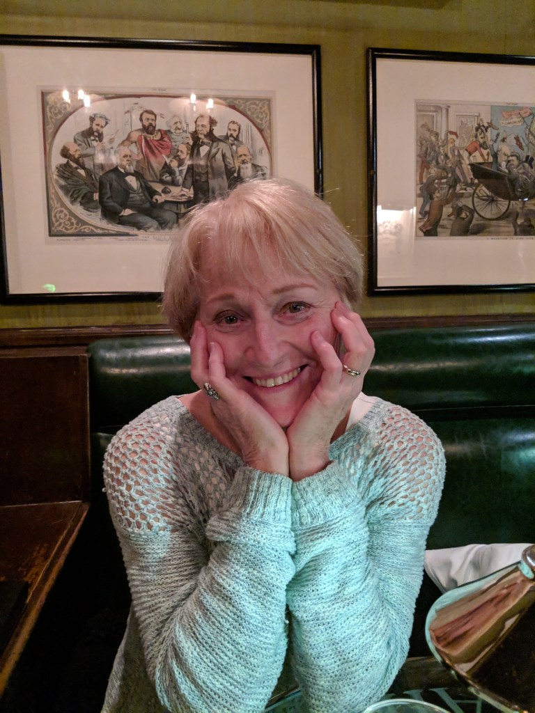 A woman with short blonde hair smiling and resting her chin on her hands, seated in a cozy restaurant with framed artworks on the walls.