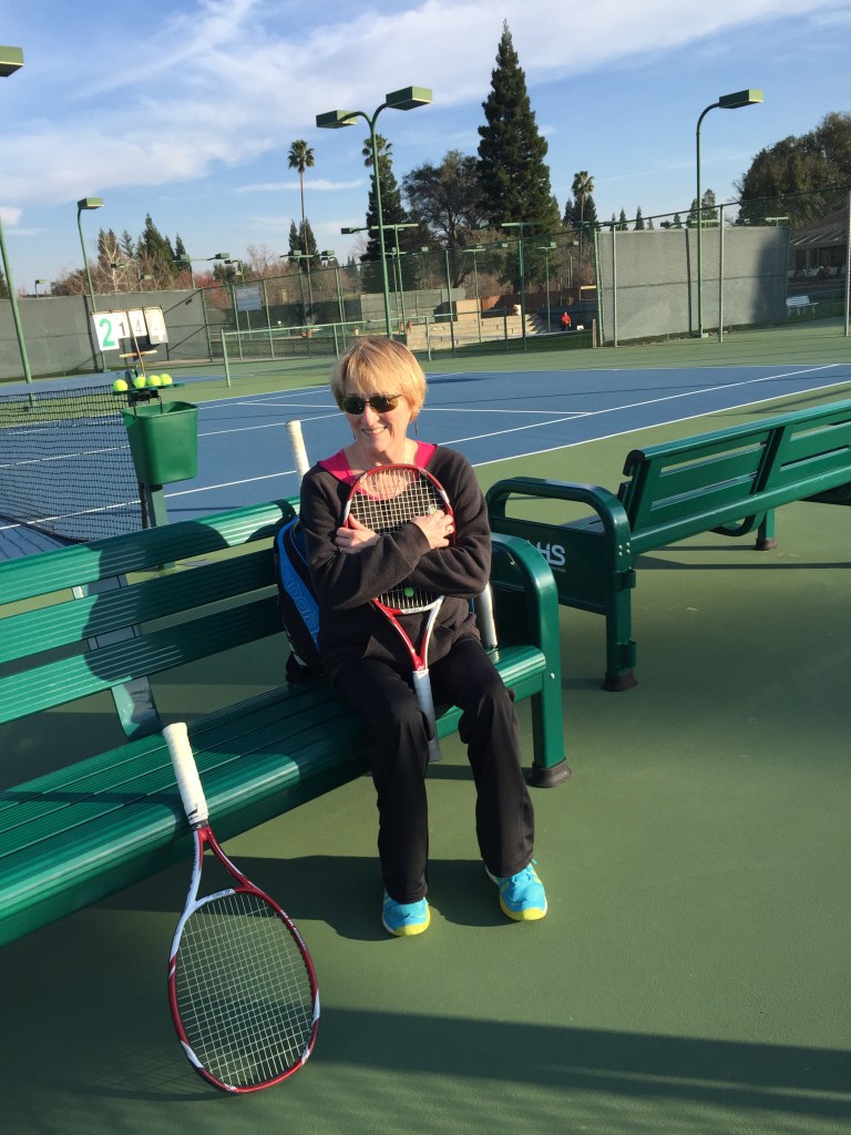 A woman sitting on a bench at a tennis court, holding a tennis racket and smiling. In the background, there are multiple tennis courts with green fences and palm trees.