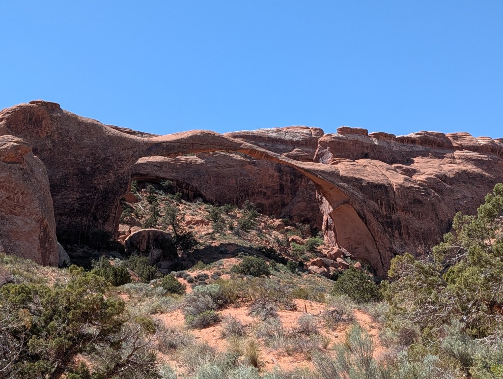 A scenic view of a natural stone arch nestled between rugged red rock formations, with sparse vegetation in the foreground against a clear blue sky.