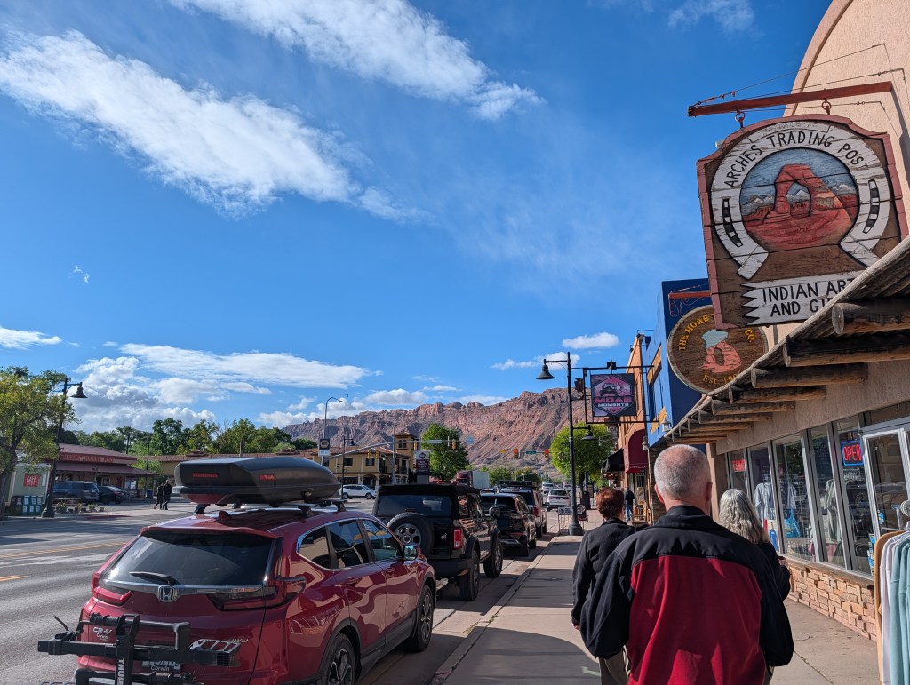 A street view of a small town with shops, including a sign for Arches Trading Post. People walk along the sidewalk, and cars are parked nearby. The sky is bright with clouds and there are mountains in the background.