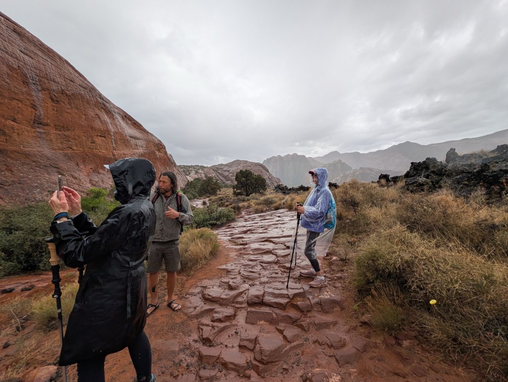 Three hikers on a wet, rocky trail in a desert landscape, wearing rain gear amidst cloudy skies.