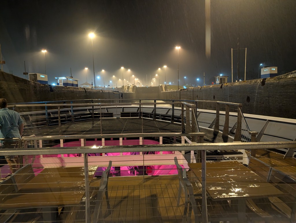 View from the deck of a boat in a lock at night, with rain visible and illuminated structures along the waterway.