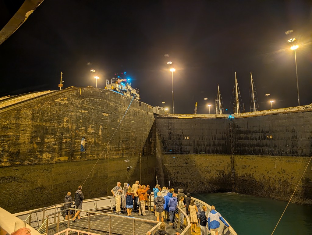 A group of people gathered at night near a large lock structure with illuminated ships in the background.