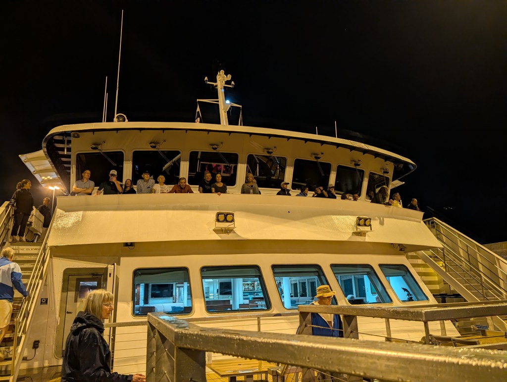 A nighttime view of a ferry boat with passengers on the upper deck, preparing to depart.