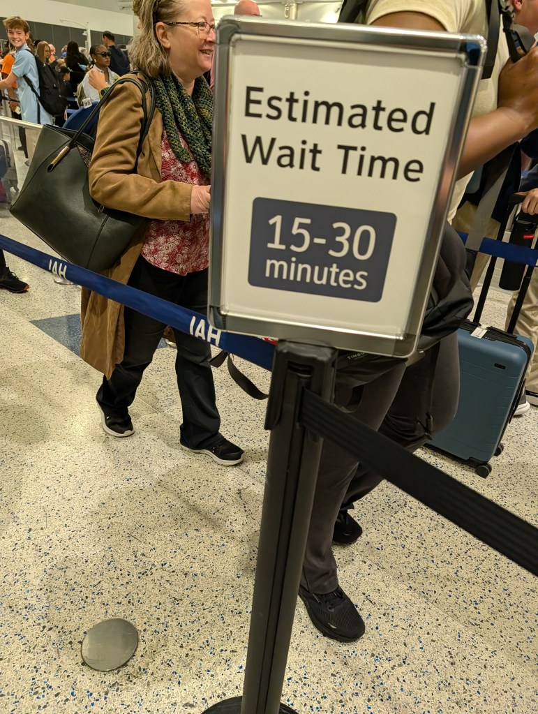 A sign displaying an estimated wait time of 15-30 minutes at an airport queue with people in line.