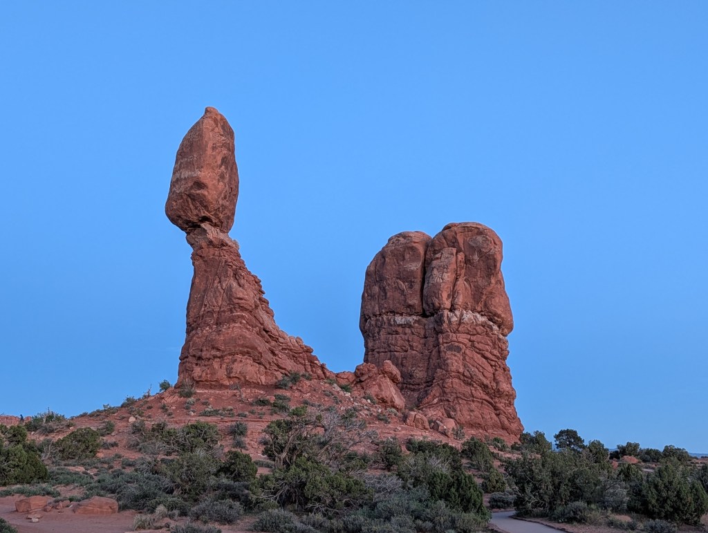 A balanced rock formation in a desert landscape with blue sky, featuring a tall, slender column topped by a large boulder.