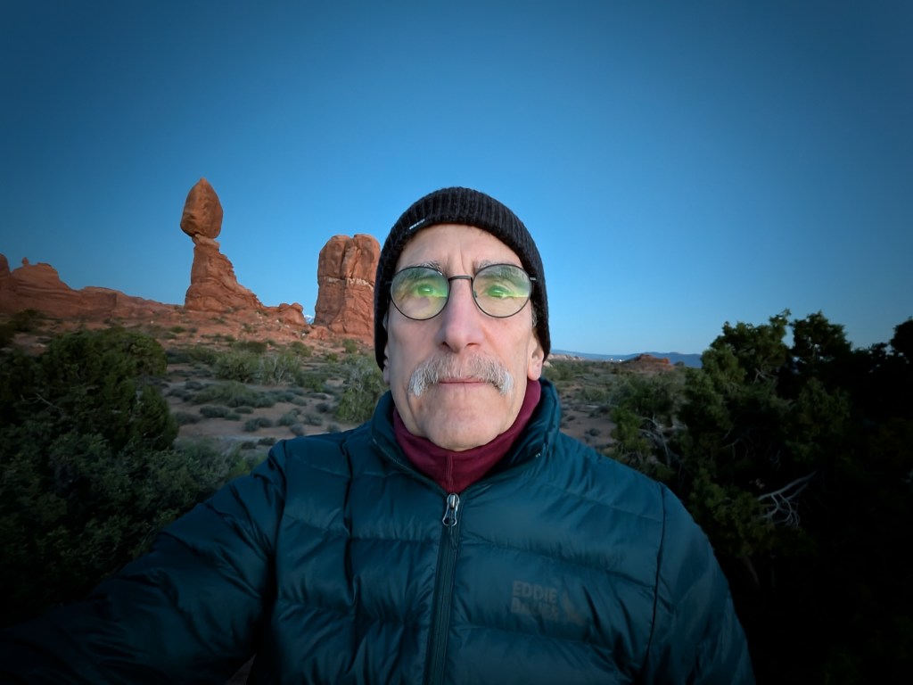 A man with glasses and a winter hat stands in front of distinctive rock formations during dawn, surrounded by desert vegetation.
