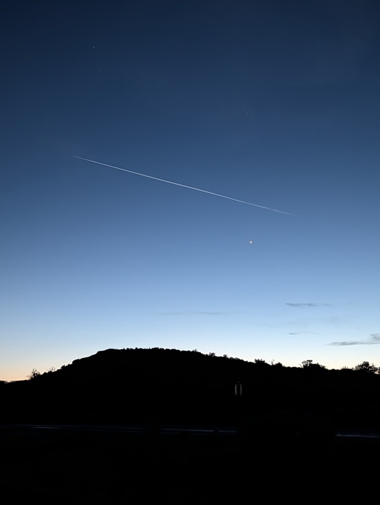 A clear evening sky transitioning from dusk to night, featuring a prominent contrail from an aircraft and a small celestial body, with a silhouetted mountain range in the foreground.