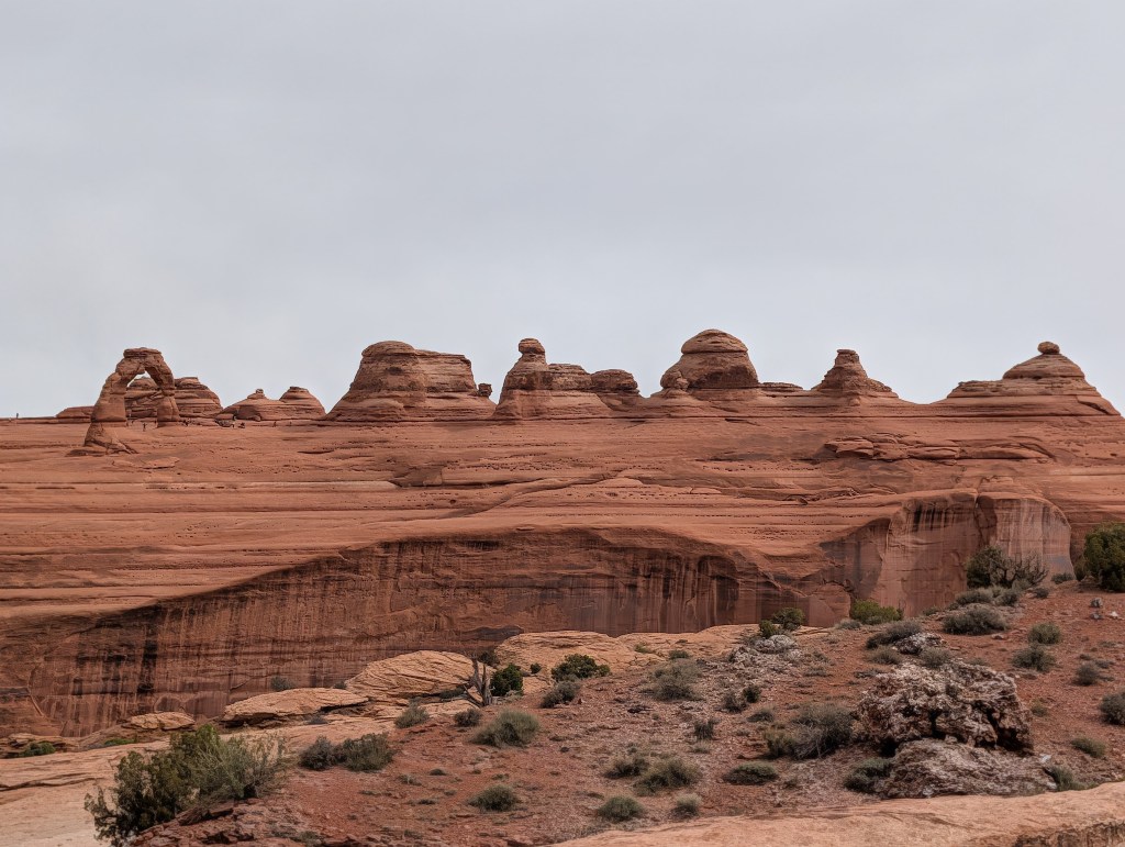 A landscape view of reddish rock formations under a cloudy sky, featuring distinct hoodoo shapes and cliffs.