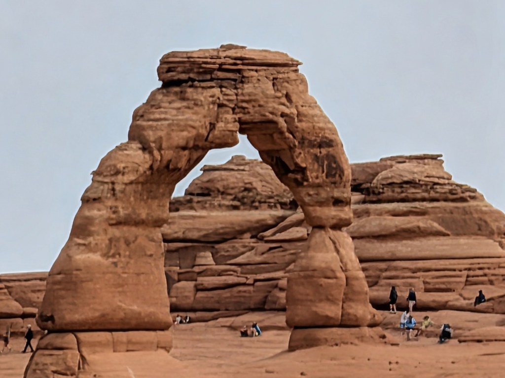 A natural stone arch formation with visitors in the foreground, set against a clear sky.