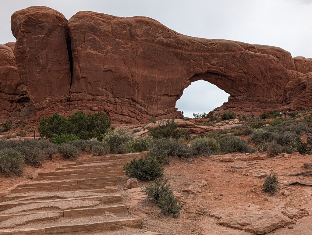 A natural rock arch formation in a red desert landscape, with a winding stone path leading towards it and sparse greenery surrounding the area.