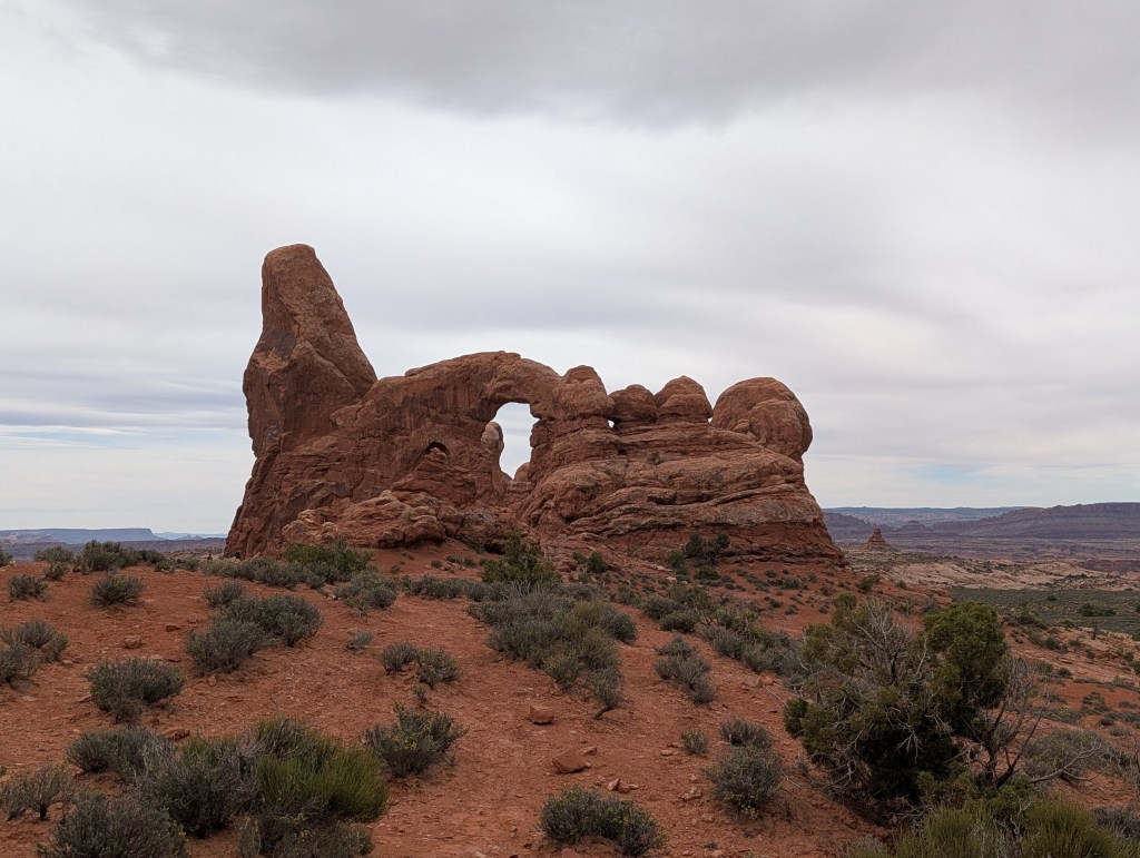 Rock formation with an arch and tower-like structure surrounded by desert terrain and shrubs.