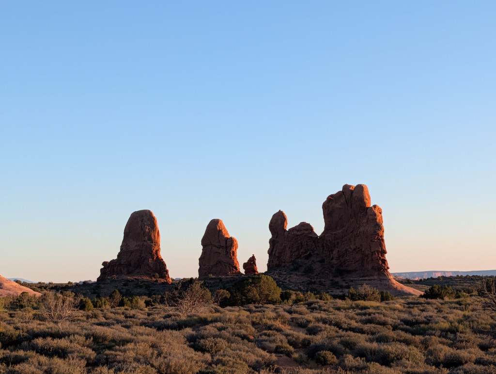A scenic view of tall red rock formations against a clear blue sky, surrounded by sparse vegetation.