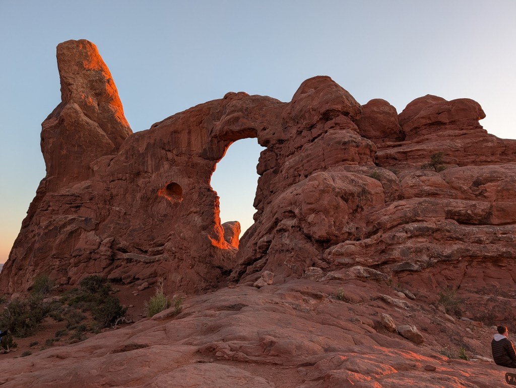A scenic view of a red rock formation with a natural arch at sunset, showcasing the vibrant colors of the rock and the clear sky.