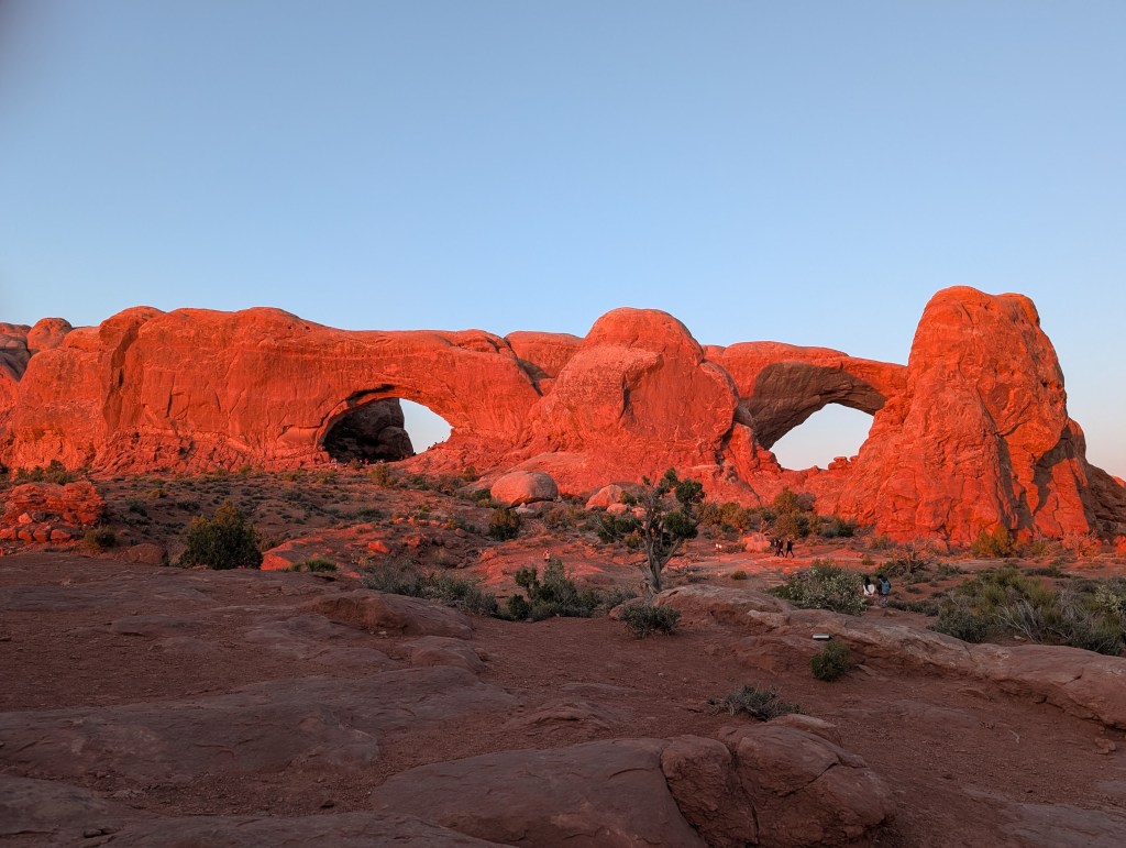 Sunset view of arches in a desert landscape, showcasing vibrant red rock formations and sparse vegetation.