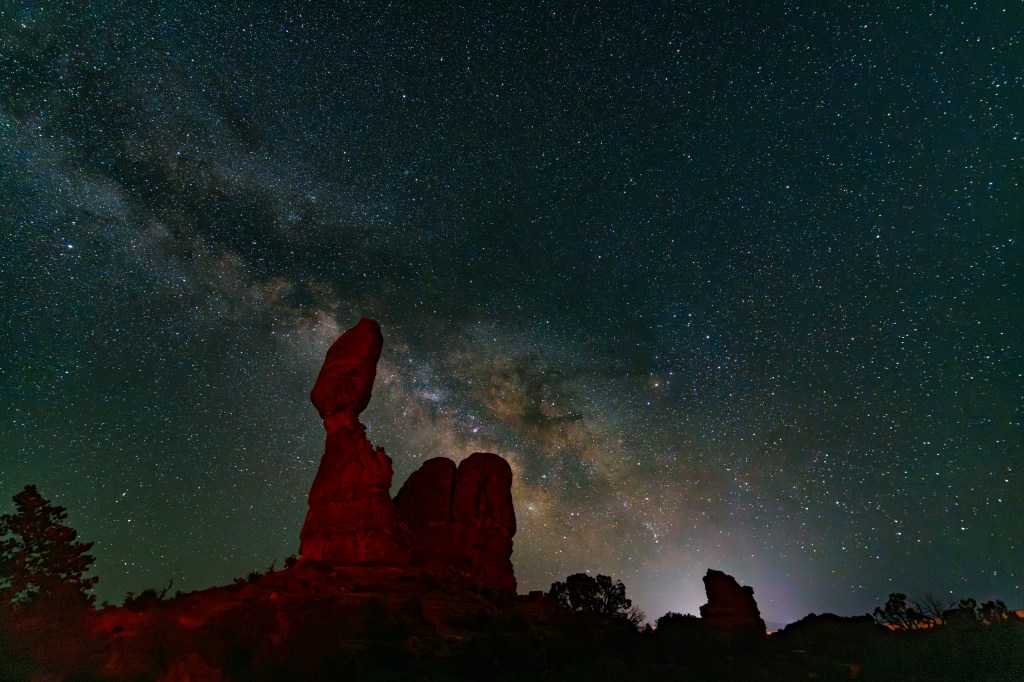 A night sky filled with stars and the Milky Way, featuring striking rock formations in the foreground illuminated by faint red light.