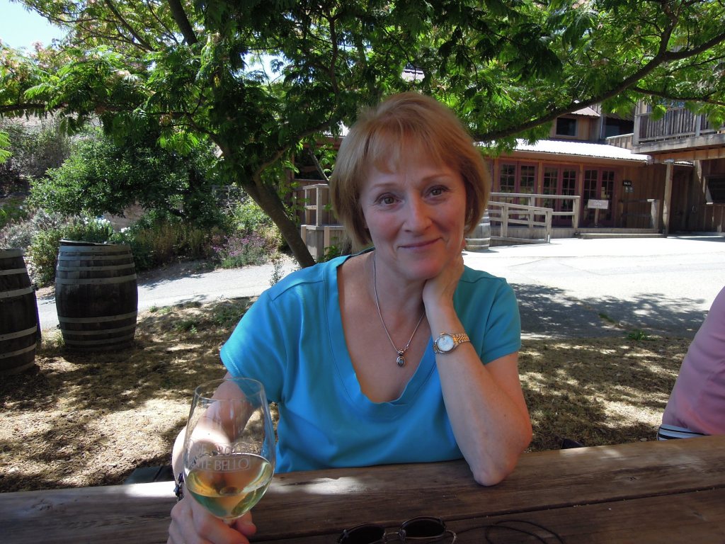 A woman sitting at an outdoor table, holding a glass of white wine, smiling and resting her chin on her hand. In the background, there are wooden barrels and a rustic building.