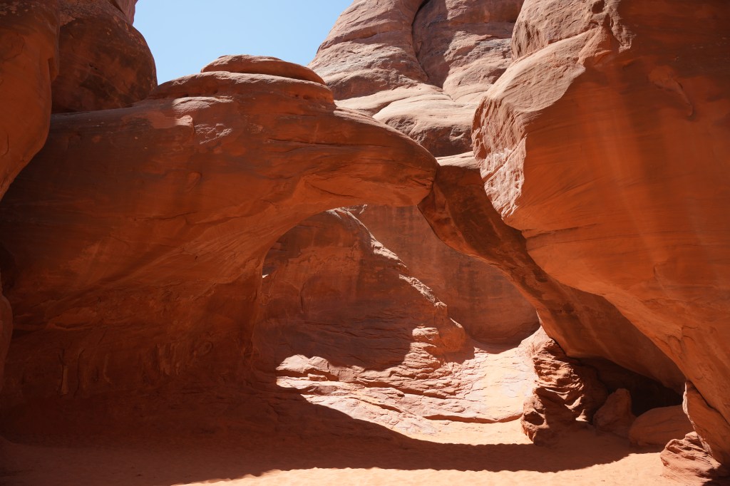 A scenic view of red rock formations in a narrow canyon, featuring a natural arch and soft sandy ground under bright blue skies.