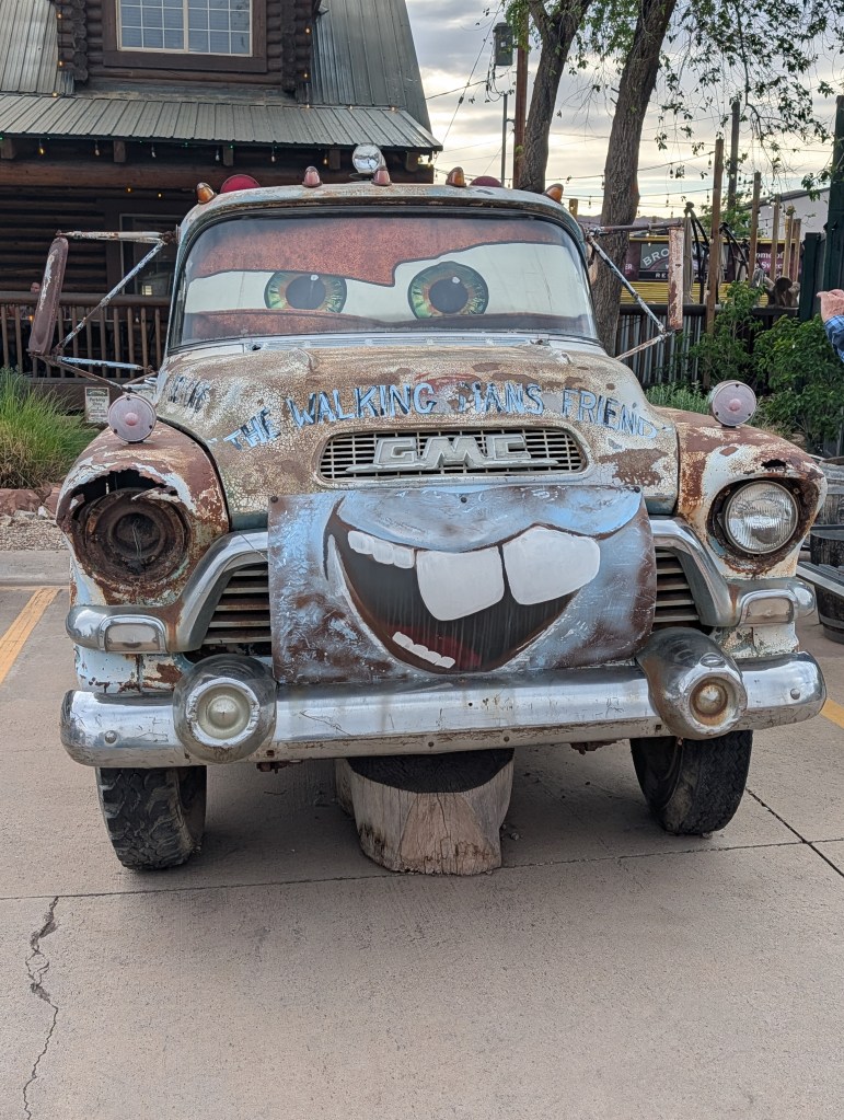 A vintage GMC truck with a playful face painted on the front, featuring large eyes and a big smiling mouth. The truck is rusted and weathered, parked in front of a wooden building with trees and a cloudy sky in the background.