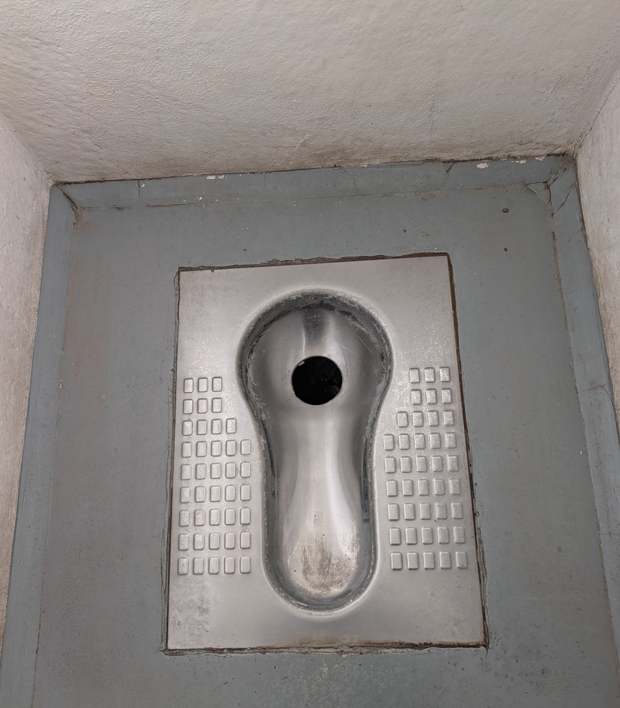 A stainless steel squat toilet in a restroom, featuring a round opening in the center and textured foot positions on either side.