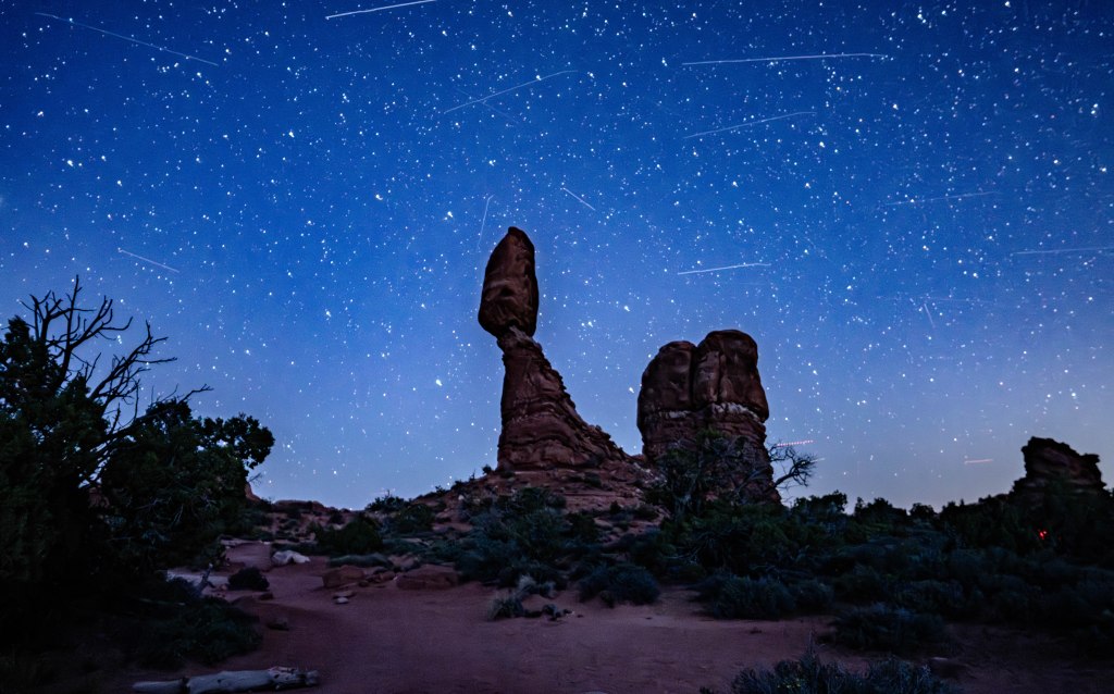 A starry night sky over a unique rock formation, featuring a prominent balancing rock and lush vegetation silhouetted against the blue background.
