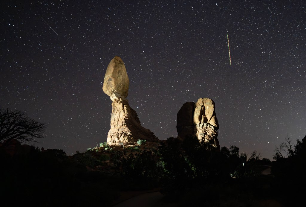 A night sky filled with stars above towering rock formations, with a prominent balancing rock illuminated by light, and a faint streak of what appears to be a meteor or satellite.
