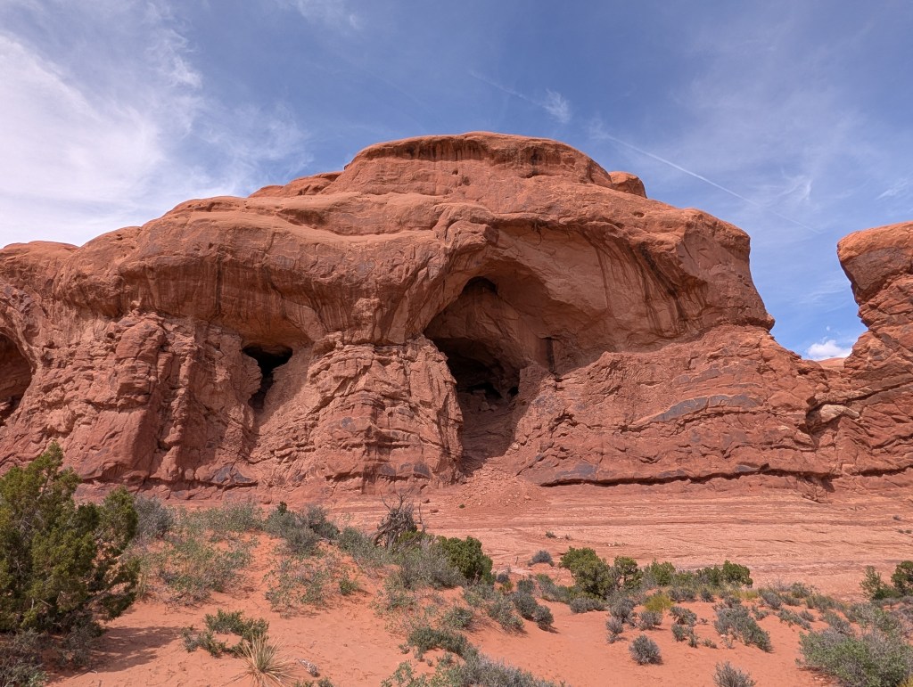 A large red rock formation with a prominent cave-like opening, surrounded by desert vegetation and a clear blue sky.