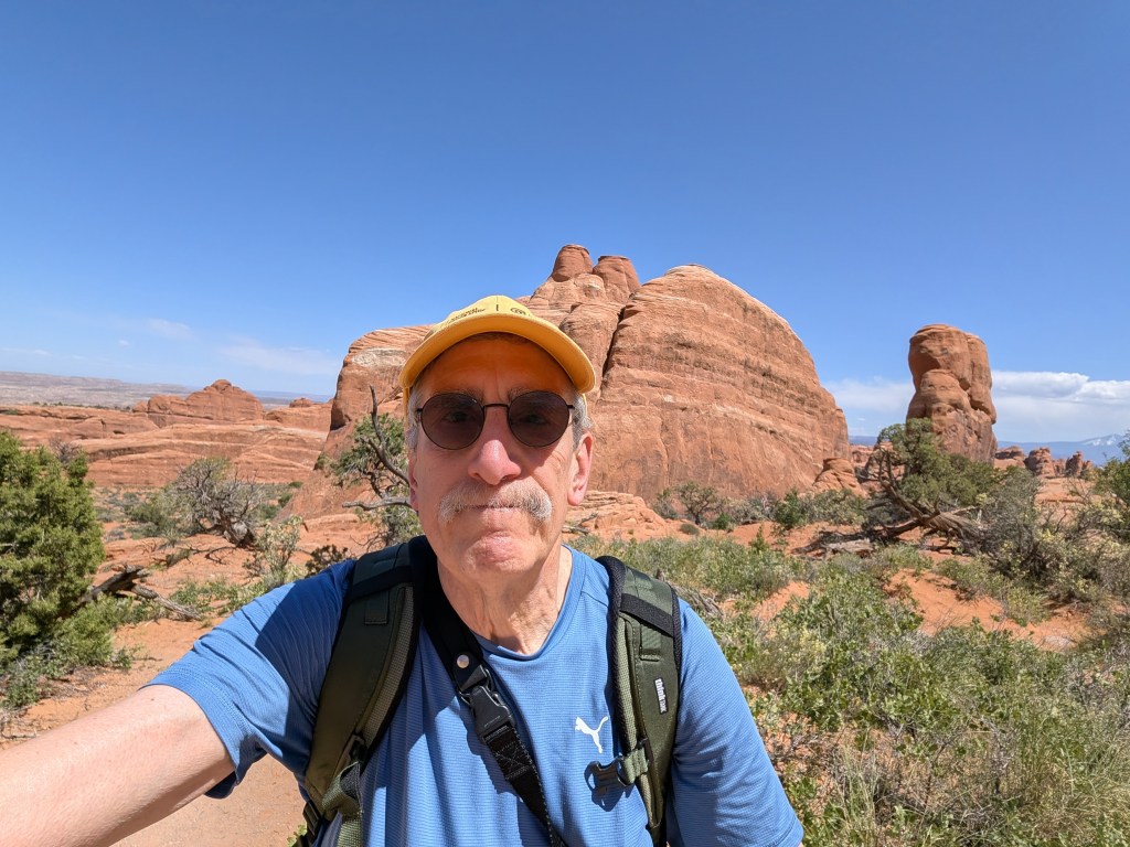 A person wearing sunglasses and a yellow cap stands in front of reddish rock formations in a desert landscape under a clear blue sky.