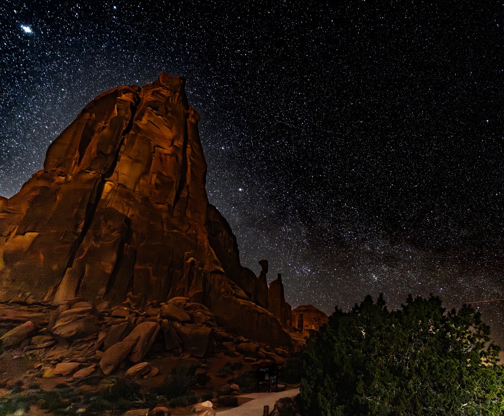A night sky filled with stars above a rocky landscape, featuring illuminated rock formations and greenery in the foreground.