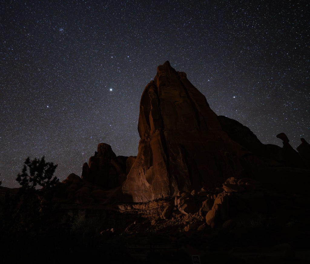 A dark night sky filled with stars above a large, illuminated rock formation.