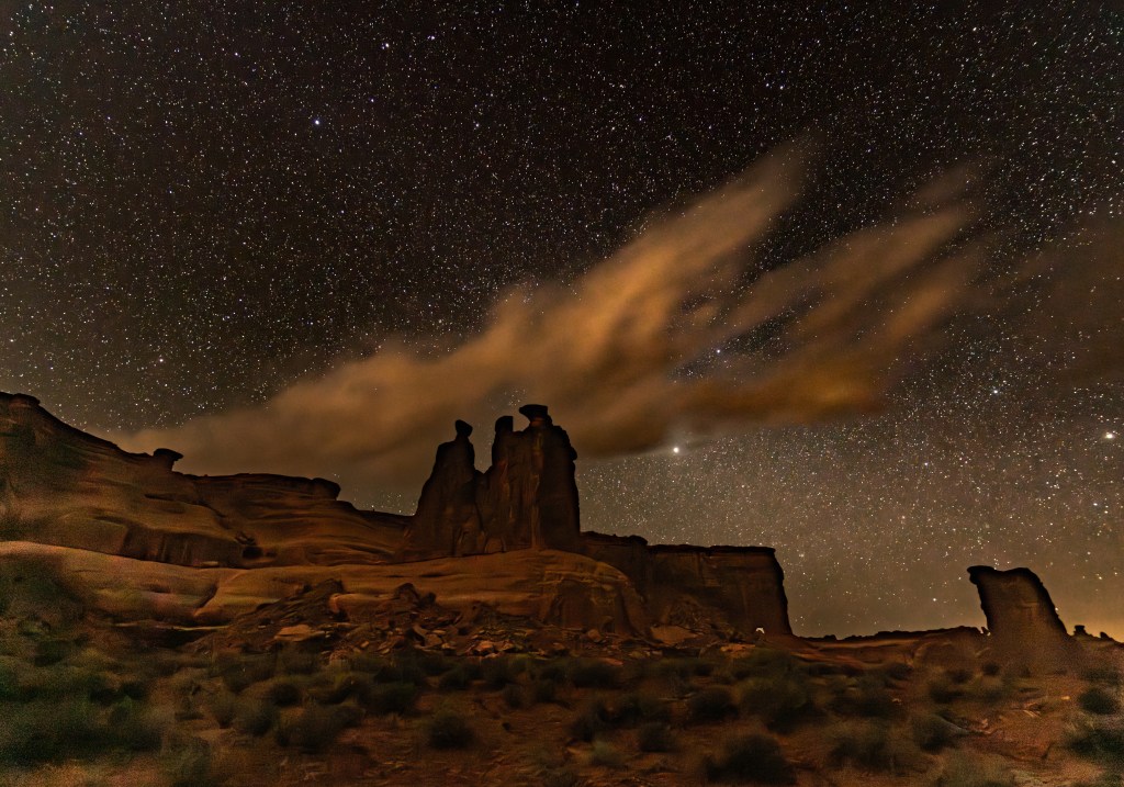 Starry night sky over rock formations with clouds illuminated by moonlight.