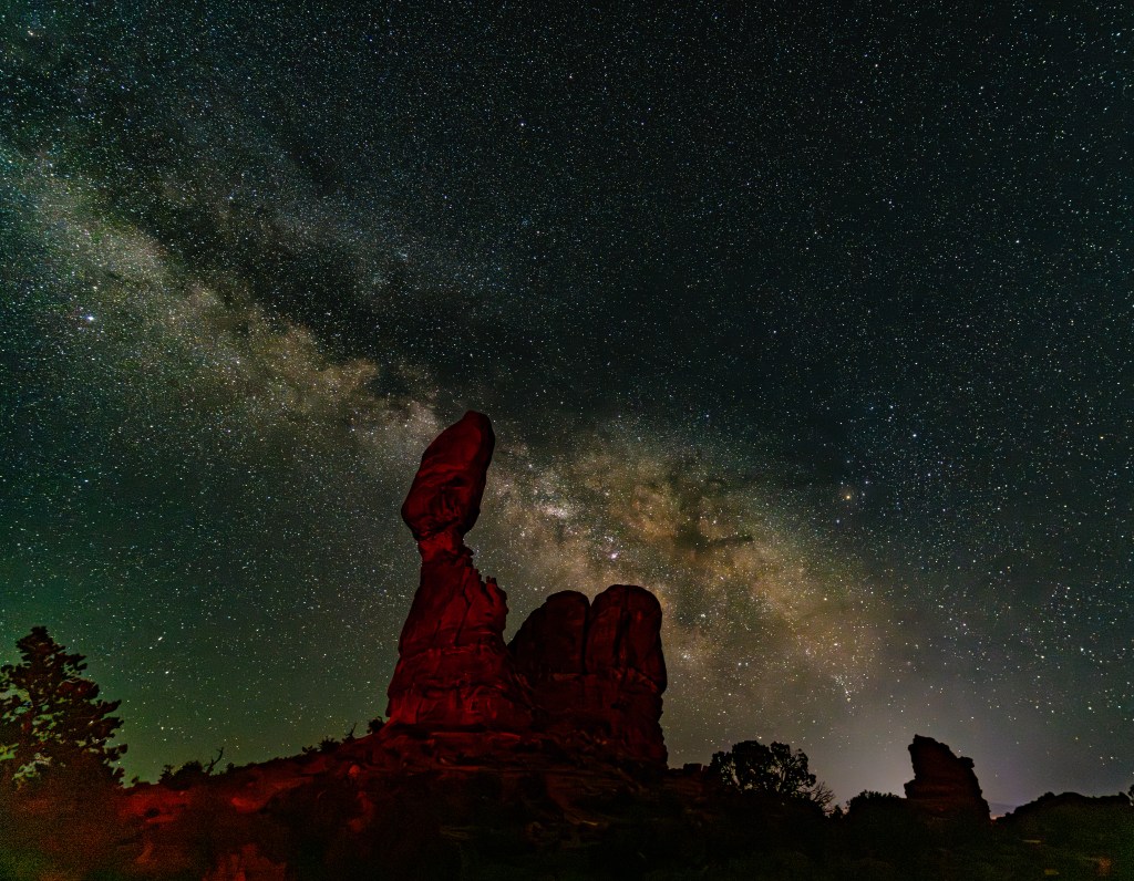Night sky with the Milky Way visible above unique rock formations, illuminated by red light.