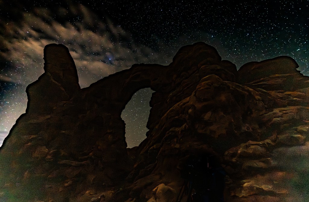 Silhouette of a rock arch against a starry night sky with clouds.