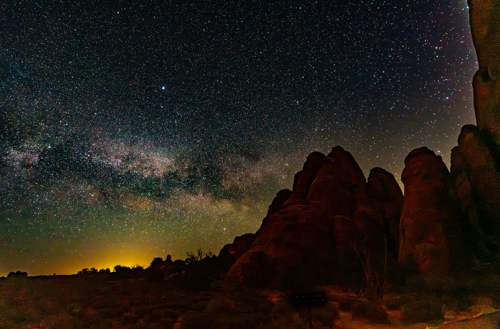 A starry night sky featuring the Milky Way, above rocky formations and a faint horizon glow.