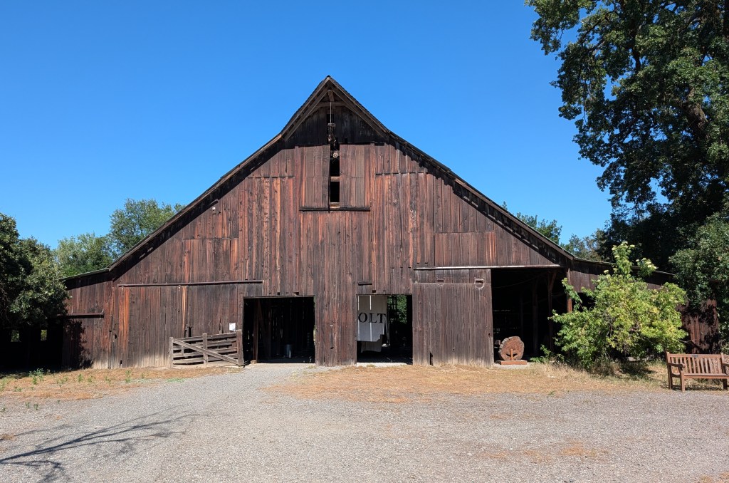 Front view of a large rustic barn with a pointed roof, surrounded by trees and a gravel path.