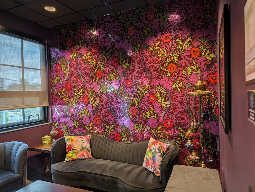 A cozy seating area featuring a dark sofa with colorful floral cushions, a chair, and a decorative side table, set against a vibrant floral-patterned wallpaper in shades of purple, red, and green.