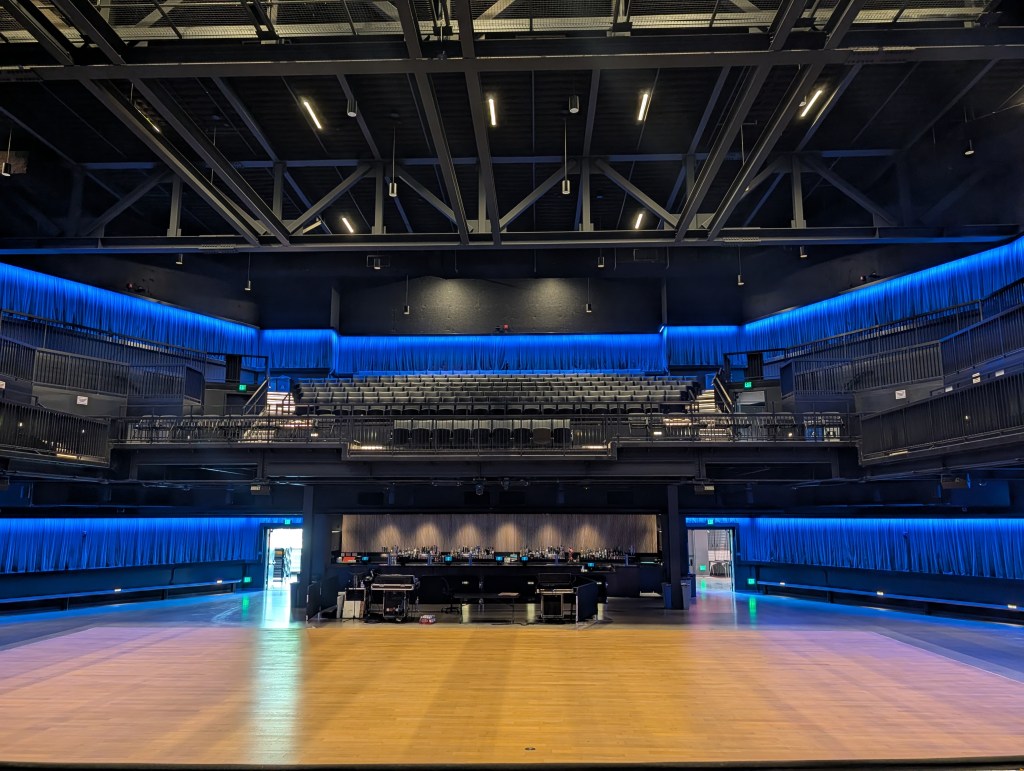 View of an empty performance stage with a wooden floor, featuring blue lighting along the upper walls and seating areas.