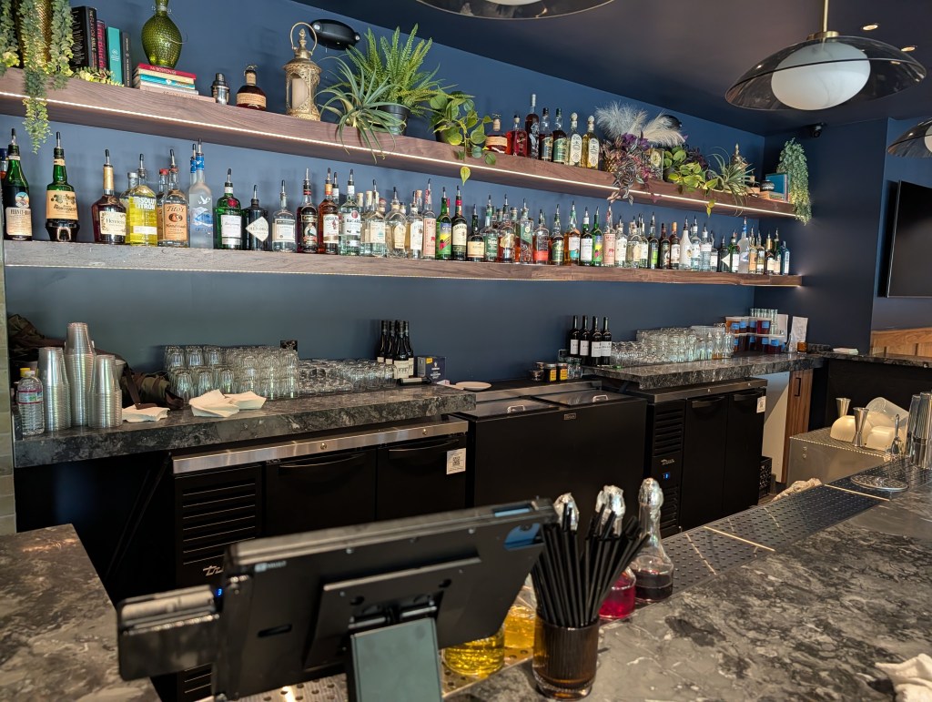 A well-stocked bar with various bottles of liquor displayed on shelves, along with glassware and a cash register on the counter.