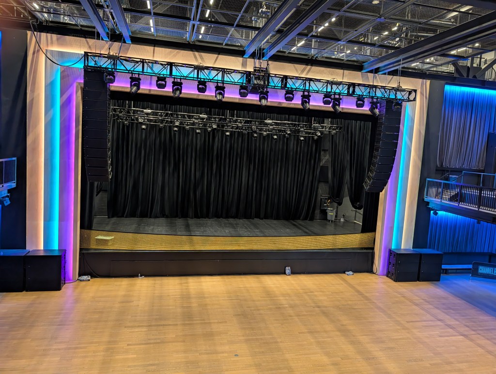 A spacious stage setup with black curtains and professional stage lighting, featuring a wooden floor and colored ambient lighting along the walls.