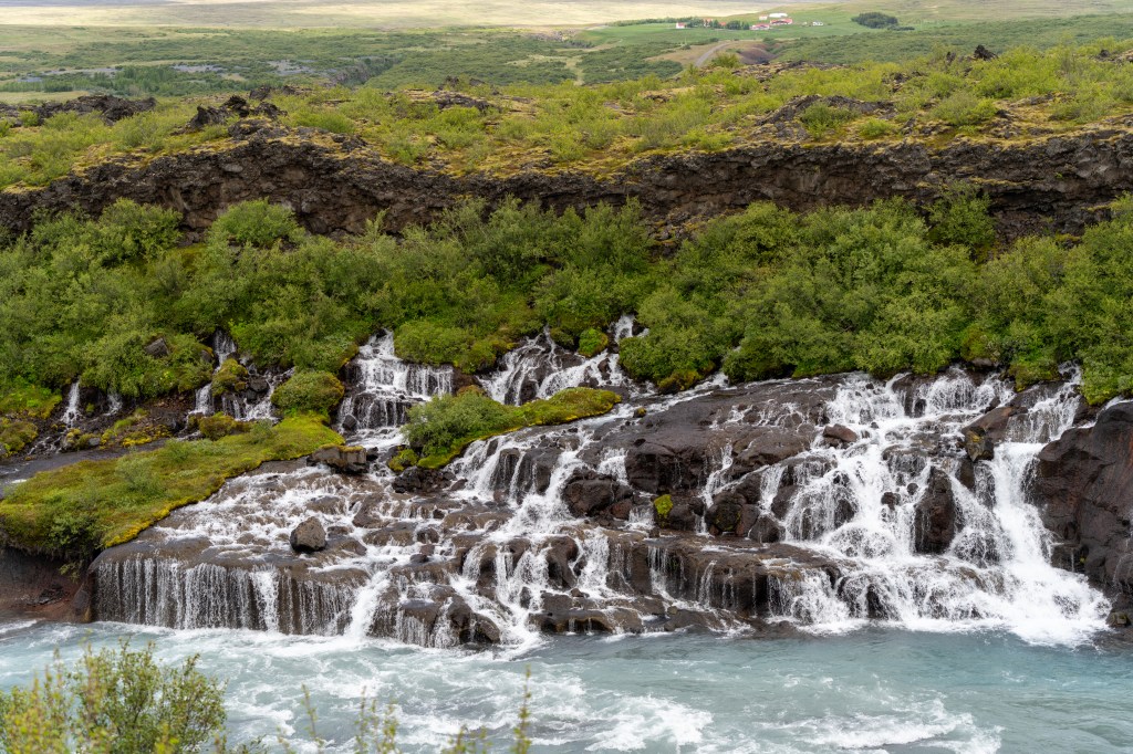 A scenic view of a cascading waterfall over rocky terrain, surrounded by lush green vegetation and a distant landscape with rolling hills.
