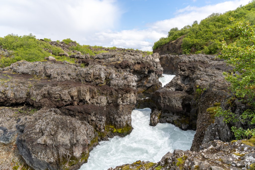 A rocky canyon with rushing water flowing through it, surrounded by greenery and under a partly cloudy sky.