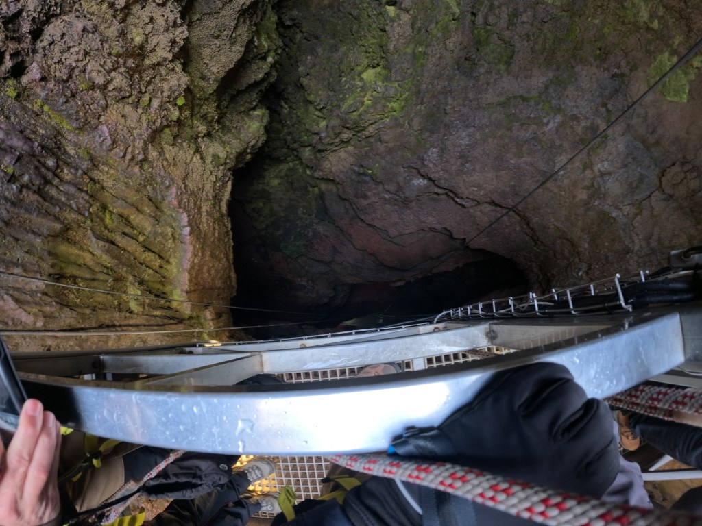 View looking down into a deep cave or pit, showing rocky walls and a metal ladder leading down.